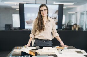 Christelle Dietsch stands at a studio table covered with fabric and material samples, with a cabin interior design displayed on a screen behind her.