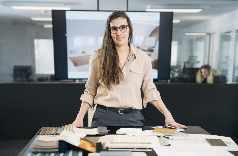 Christelle Dietsch stands at a studio table covered with fabric and material samples, with a cabin interior design displayed on a screen behind her.