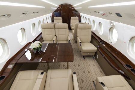 The interior of a business jet featuring cream leather club seating arranged around a dark wood table topped with a white floral arrangement. The cabin has white overhead panels with recessed lighting, rich dark wood veneer accents, and a geometric-patterned beige/cream carpet.