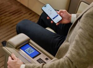 A passenger using a smartphone alongside an armrest-mounted touchscreen displaying a QR code, inside a business aircraft cabin.
