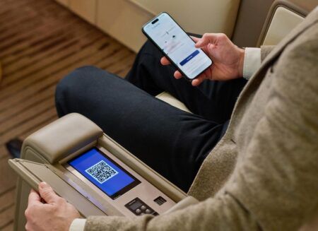 A passenger using a smartphone alongside an armrest-mounted touchscreen displaying a QR code, inside a business aircraft cabin.
