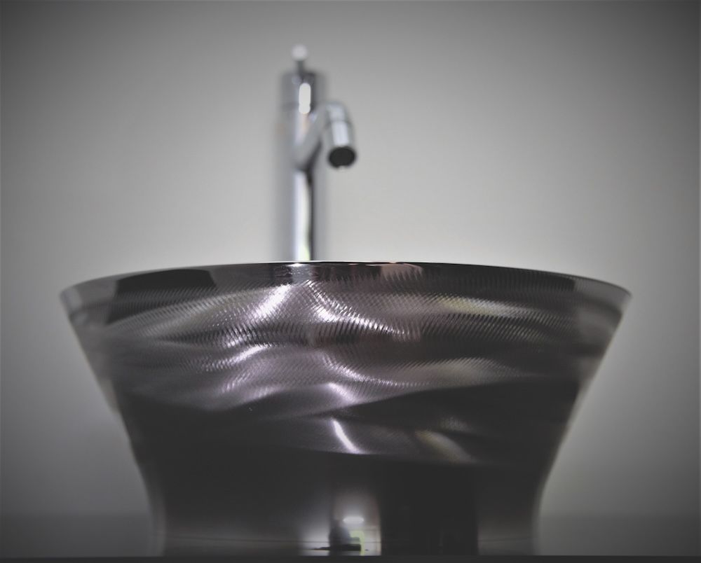 Photograph of a polished dark stainless steel vessel sink with undulating wave-texture surface finish and a chrome tap, against a neutral grey background.
