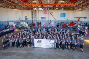 Large group of Textron Aviation staff in matching grey shirts standing in a hangar with two aircraft and red, white and blue balloon displays, holding a banner marking 45 years of the Tampa service centre