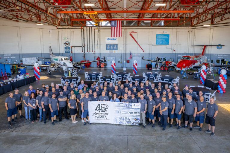Large group of Textron Aviation staff in matching grey shirts standing in a hangar with two aircraft and red, white and blue balloon displays, holding a banner marking 45 years of the Tampa service centre