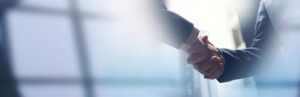 Stock photograph of two people in business suits shaking hands, with a soft-focus office window in the background, used to illustrate a corporate acquisition agreement.