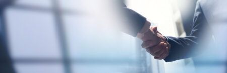 Stock photograph of two people in business suits shaking hands, with a soft-focus office window in the background, used to illustrate a corporate acquisition agreement.