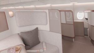 Close-up render of a single premium-class shell seat, upholstered in textured light grey fabric with a charcoal cushion. The suite is enclosed by a tall padded privacy screen trimmed in a rose gold finish - with a frosted upper section. A cylindrical reading light is mounted by the head rest. A tray bearing a pink drink and a small dish is partially visible in the foreground. Additional suites are visible beyond the partition.