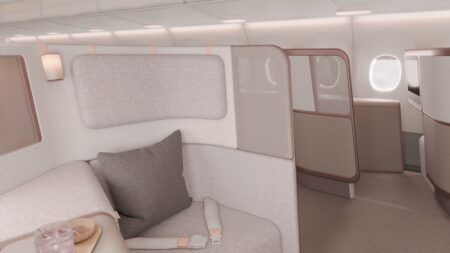 Close-up render of a single premium-class shell seat, upholstered in textured light grey fabric with a charcoal cushion. The suite is enclosed by a tall padded privacy screen trimmed in a rose gold finish - with a frosted upper section. A cylindrical reading light is mounted by the head rest. A tray bearing a pink drink and a small dish is partially visible in the foreground. Additional suites are visible beyond the partition.