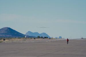 Photograph of a long, slender white unmanned glider-style aircraft flying low over a concrete airfield apron, with mountain ridges in the background and a person in a red jacket watching from the runway