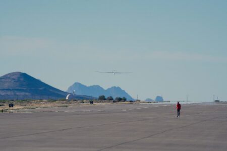 Photograph of a long, slender white unmanned glider-style aircraft flying low over a concrete airfield apron, with mountain ridges in the background and a person in a red jacket watching from the runway