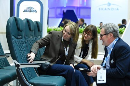 Photograph of three attendees inspecting a row of green leather Acro aircraft seats at an exhibition stand, with branded backdrops visible behind them