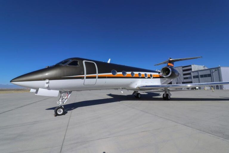 Photograph of a Gulfstream GIV business jet on a tarmac, painted white on the lower fuselage with a black upper section and an orange accent stripe running the length of the aircraft, parked outside a hangar under a clear blue sky
