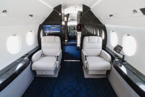 Interior of refurbished Dassault Falcon 2000EXy showing two white leather club seats facing each other, a forward bulkhead monitor displaying a world map, and dark wood veneer panelling with blue patterned carpet