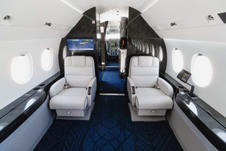 VIP Completions delivers refurbished Dassault Falcon 2000EX EASy Interior of refurbished Dassault Falcon 2000EXy showing two white leather club seats facing each other, a forward bulkhead monitor displaying a world map, and dark wood veneer panelling with blue patterned carpet