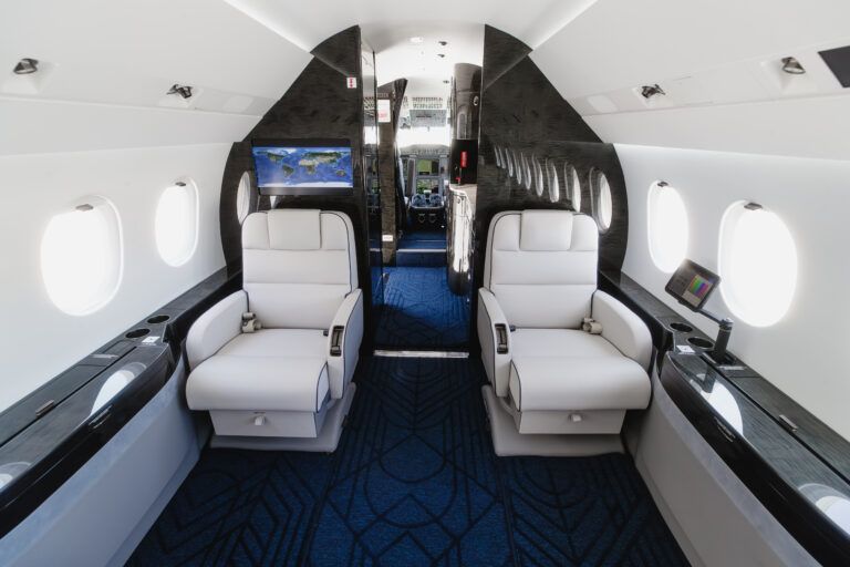 VIP Completions delivers refurbished Dassault Falcon 2000EX EASy Interior of refurbished Dassault Falcon 2000EXy showing two white leather club seats facing each other, a forward bulkhead monitor displaying a world map, and dark wood veneer panelling with blue patterned carpet