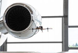 A four-rotor inspection drone hovers next to the polished engine nacelle of a business jet inside a hangar, with the engine fan blades visible behind it