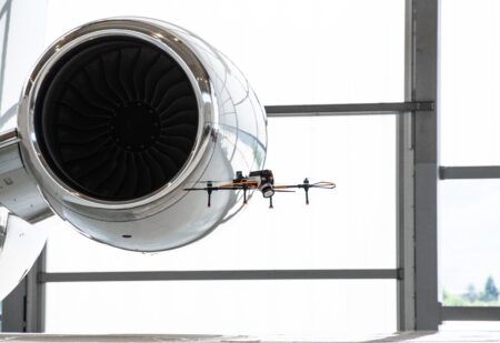 Jet Aviation launches inspections by automated drone and AI in the US A four-rotor inspection drone hovers next to the polished engine nacelle of a business jet inside a hangar, with the engine fan blades visible behind it
