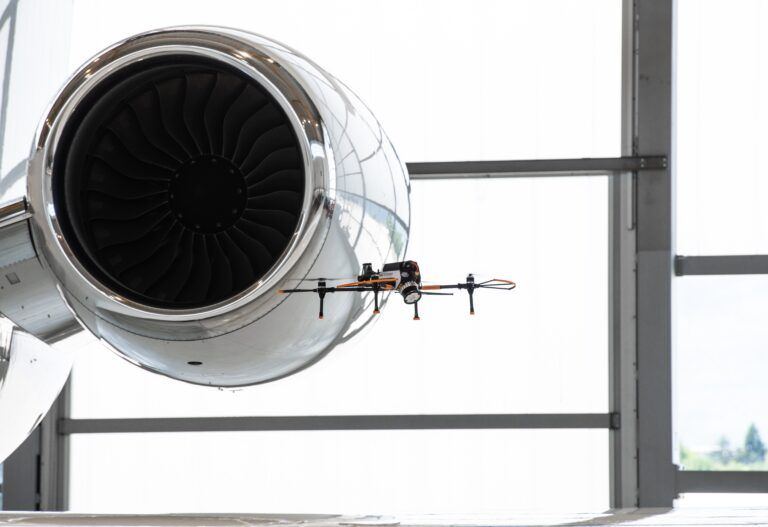 A four-rotor inspection drone hovers next to the polished engine nacelle of a business jet inside a hangar, with the engine fan blades visible behind it