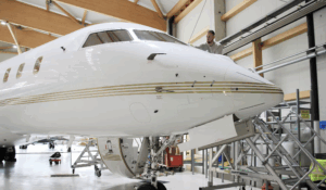 Photograph of a white Bombardier Global 5000 business jet parked on a tarmac apron with its airstair door open, shown in side profile against a clear background.