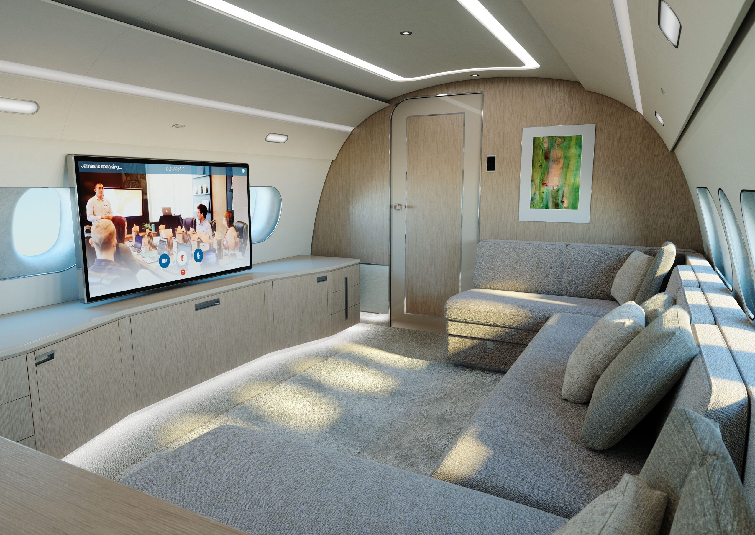 Render of the ACJ TwoTwenty guest lounge area showing a large wall-mounted screen displaying a video conference, a grey corner sofa with cushions, soft lighting and a framed artwork on a wood-panelled bulkhead