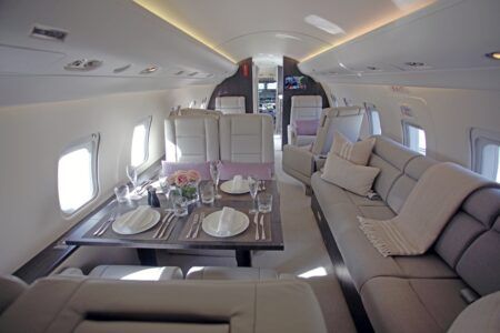 Photograph of the interior cabin of a Bombardier Challenger 604 business jet, showing cream leather club seats, a grey three-seat divan with cushions and throw, a set dining table with china, glassware and pink roses, and a flat-screen monitor toward the forward cabin.
