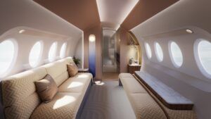 A bright, daytime render of a private jet cabin lounge area. On the left bulkhead is the ‘floating orb’ light feature, a tall panel gradually turning from a natural daylight colour at the top to deep blue at the bottom. The orb, representing the sun, is at the top, and the cabin is lit in a soft, natural daytime ambiance. A divan on the left is upholstered in a cream herringbone fabric, also used for bench seating on the right, which has a grey-veined stone surface element. In front of the right bulkhead is a warm-lit bar niche containing stemware and a silver champagne service. The ceiling is gently curved and features recessed lighting.