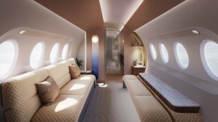 FEATURE: JPA Design on circadian lighting A bright, daytime render of a private jet cabin lounge area. On the left bulkhead is the ‘floating orb’ light feature, a tall panel gradually turning from a natural daylight colour at the top to deep blue at the bottom. The orb, representing the sun, is at the top, and the cabin is lit in a soft, natural daytime ambiance. A divan on the left is upholstered in a cream herringbone fabric, also used for bench seating on the right, which has a grey-veined stone surface element. In front of the right bulkhead is a warm-lit bar niche containing stemware and a silver champagne service. The ceiling is gently curved and features recessed lighting.