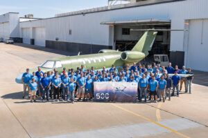 Large group of workers in blue 500 t-shirts pose in front of an unpainted green business jet outside a factory hangar