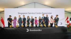 A group of officials and executives hold ceremonial golden spades at a groundbreaking event for Bombardier's Singapore Service Centre Expansion. The Singapore and Canadian flags are displayed on either side of the stage.