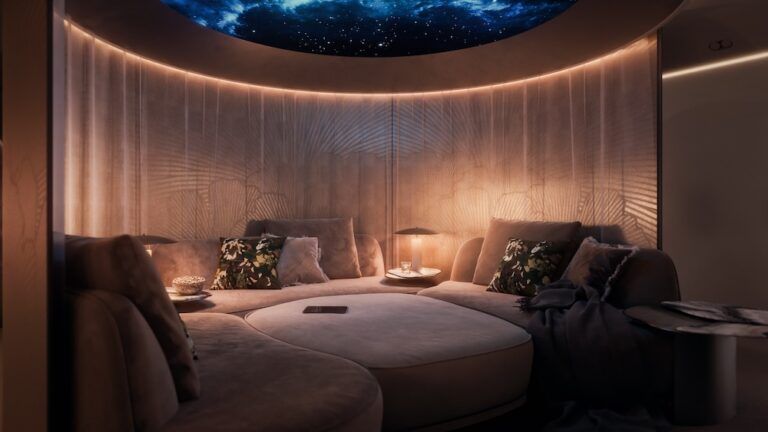 The adaptive living area configured as a cinema, with sofas arranged around an ottoman, screens around, beneath a circular overhead display showing a starfield. Warm ambient lighting.