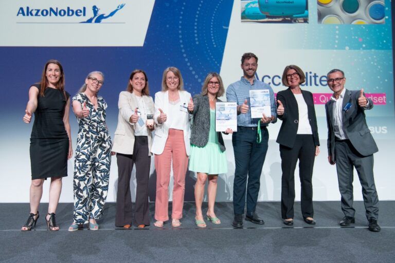 AkzoNobel team celebrates on stage at Airbus accreditation awards ceremony, giving thumbs up and holding certificates