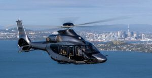 A dark grey Airbus ACH160 helicopter in flight over the water with the Auckland skyline behind