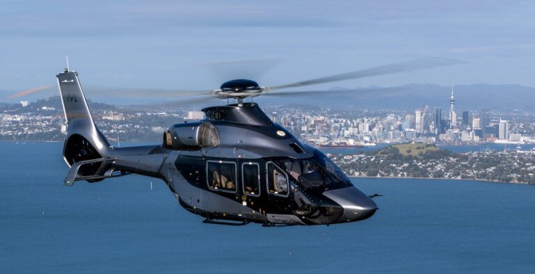 A dark grey Airbus ACH160 helicopter in flight over the water with the Auckland skyline behind