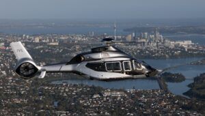 A dark grey Airbus ACH160 helicopter in flight over the water with city skyline behind