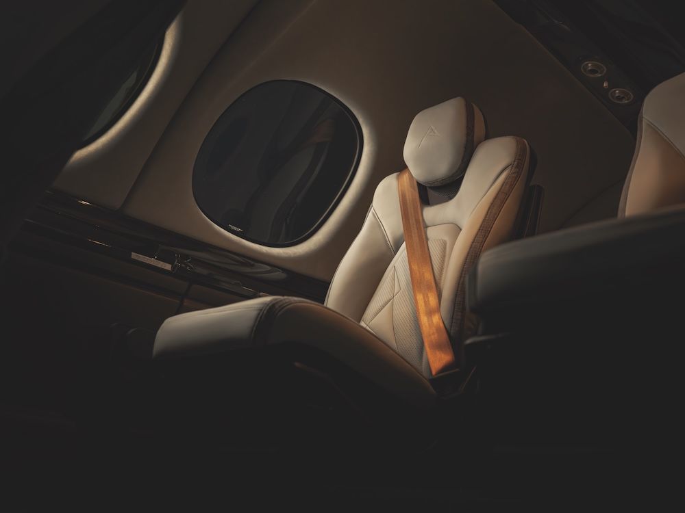 Close-up of a redesigned cream leather seat with tan seatbelt strap in the G3 Vision Jet cabin, showing an oval window and stitched headrest detail.