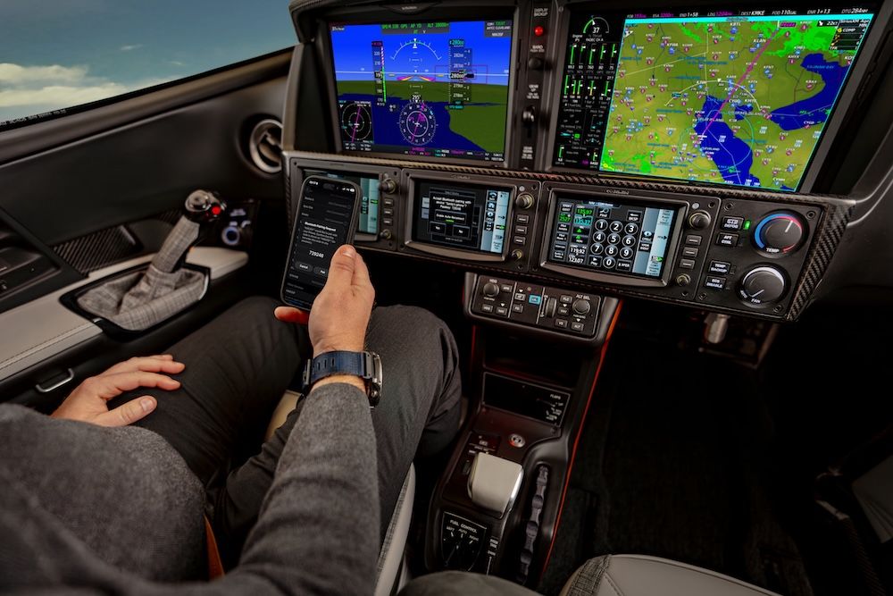 View from the pilot's seat of the G3 Vision Jet's flight deck, showing multiple digital displays with navigation maps, flight data and a carbon-fibre-trimmed instrument panel, with a pilot's hand holding a smartphone.