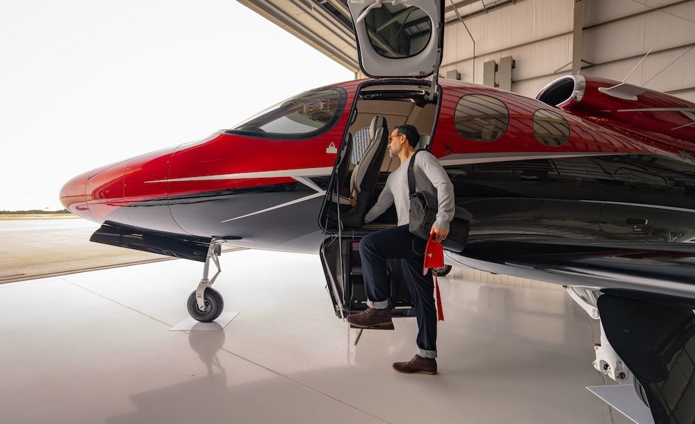A man boarding a red and black Cirrus Vision Jet parked in a hangar, stepping up through the open cabin door with a bag over his shoulder.