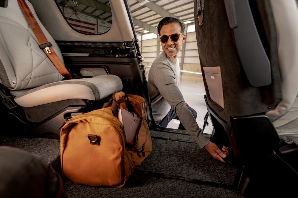 A smiling man in sunglasses crouched in the cabin doorway of the G3 Vision Jet, loading a tan duffel bag beside the redesigned passenger seats.