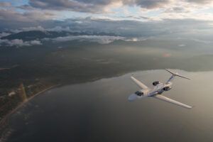 : A white light business jet banking in flight above a lake and forested coastline, with mountains and a cloudy sky in the background.