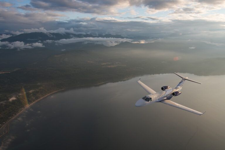 : A white light business jet banking in flight above a lake and forested coastline, with mountains and a cloudy sky in the background.