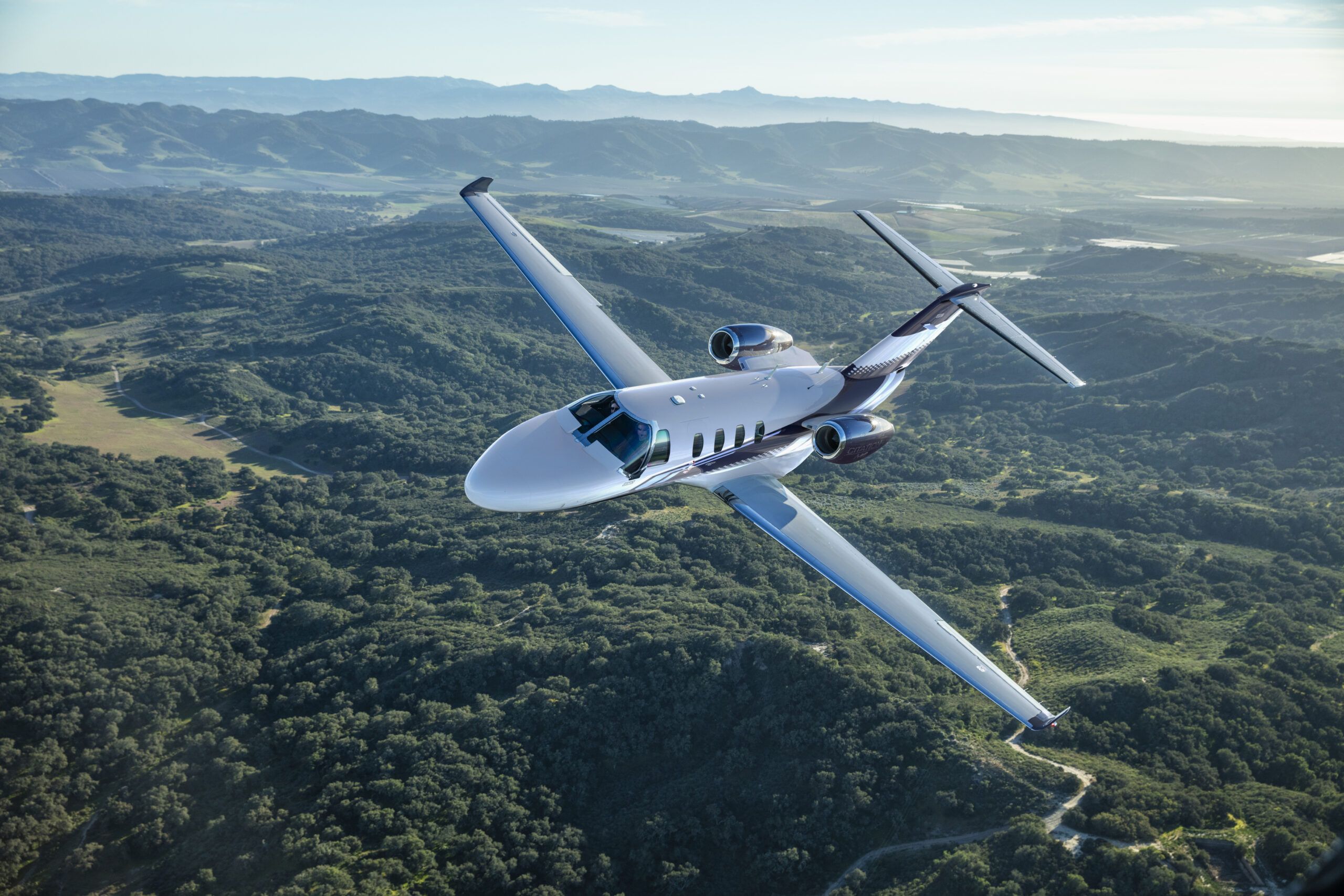A white light business jet in flight, viewed from above against terrain below.