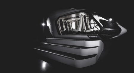 Black ACH140 helicopter mockup on a staged podium with illuminated steps, cabin interior visible through the open door showing rows of white seats, dramatic black background with two spotlights either side.