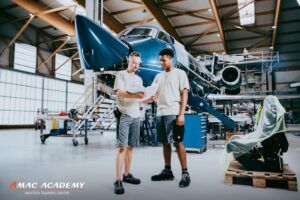 Two people in AMAC uniforms reviewing documents in front of an aircraft undergoing maintenance inside a hangar, with the AMAC Academy Aviation Training Center logo visible.