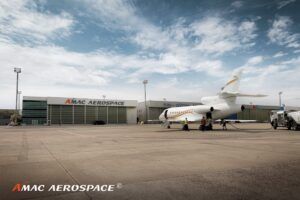A business jet on a concrete apron next to a fuel truck, with ground crew nearby and an AMAC Aerospace hangar visible in the background.