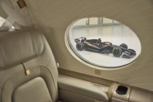 Interior of a Flexjet business jet cabin showing a beige leather seat beside an oval window, through which a black Formula 1 car is visible in a hangar.
