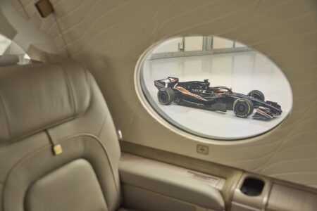 Interior of a Flexjet business jet cabin showing a beige leather seat beside an oval window, through which a black Formula 1 car is visible in a hangar.