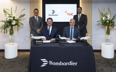 Four men at a signing ceremony, two seated and signing documents at a Bombardier-branded table, two standing behind, with three business-jet scale models on the table and a screen showing the Bombardier and THC logos