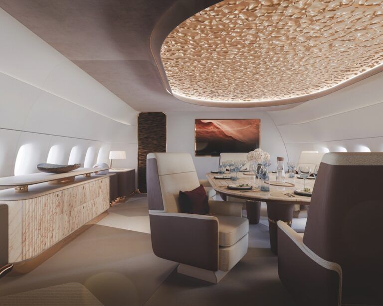 Business jet cabin interior featuring a circular dining suite with cream leather swivel chairs, a textured gold dimpled ceiling panel with concealed lighting, and a wood-veneer credenza along the sidewall.