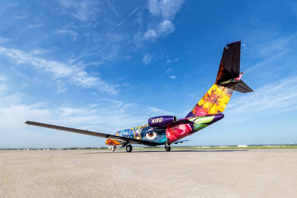 Rear three-quarter view of a business jet’s tail and engine nacelle, painted with colourful floral and eye designs, against a blue sky.