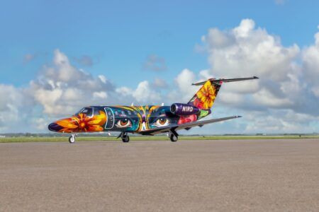 VIDEO: Duncan Aviation unveils 46-colour paint scheme Side view of a Citation CJ3+ business jet on a runway, painted in a multicoloured livery with large flowers and stylised eyes against a blue sky.